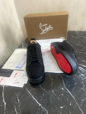 Sneakers Christian Louboutin Louis Junior Flat Suede ⚫ - Snygga svarta sneakers från Christian Louboutin i mocka med klassisk röd sula och svart broderad logga på hälen. Lågt skaft och svarta skosnören ger en clean look. Perfekta för dig som vill sticka ut med exklusiva detaljer.