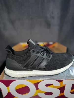 Svarta Adidas Ultraboost sneakers - Säljer ett par  helt nya svarta Adidas Ultraboost sneakers med stickad ovandel och vita Boost-sulor. Skorna har klassiska tre ränder på sidan, svart hälkappa och svart snörning. Perfekta för dig som gillar stilrena och bekväma sneakers.