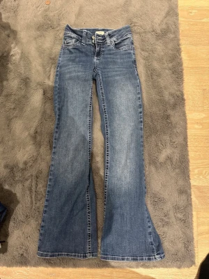 Blå bootcut jeans med detaljer - Säljer ett par blå bootcut jeans från Nelly med snygg slitning och dekorativ söm i vitt på bakfickan. Stl 32