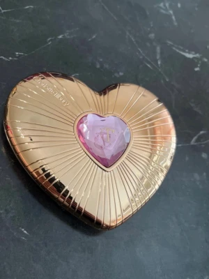 Hjärtformat rouge Charlotte Tilbury - Rouge i hjärtformad guldig dosa med lila detalj från Charlotte Tilbury. Innehåller två nyanser: en ljusrosa och en djupare rosa, båda med små hjärtmönster. Perfekt för att skapa en fräsch och romantisk look.