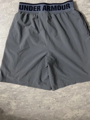 Grå träningsshorts Under Armour - Snygga grå träningsshorts från Under Armour med elastisk midja och logga på linningen. Tillverkade i ett lätt och snabbtorkande material, perfekta för gymmet eller löprundan. Klassisk passform och diskret logga på benet.