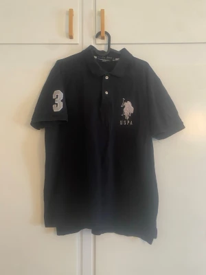 Svart pikétröja från US POLo - Klassisk svart pikétröja från US POLO med korta ärmar, krage och knappar framtill. På bröstet finns USPA-logga i vitt och på ena ärmen en stor siffra 3. Perfekt till en avslappnad men ändå snygg look.