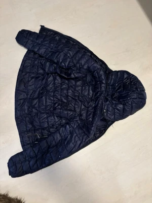 dunjacka  - Snygg mörkblå dunjacka med quiltat mönster och fast huva. Jackan har dragkedja framtill och justerbar nederkant. Perfekt för kalla vinterdagar och riktigt skön att bära. Insidan är ljusgrå och jackan har praktiska fickor. En av dragkedjorna till fickorna är trasig men är man kunnig och kan sy så är det inga problem 