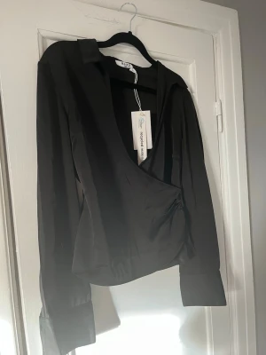 Svart omlottblus från NA-KD - Stilren svart omlottblus från NA-KD i glansigt material. Blusen har djup v-ringning, långa ärmar med breda manschetter och en markerad midja. Perfekt för dig som gillar trendiga och eleganta plagg.