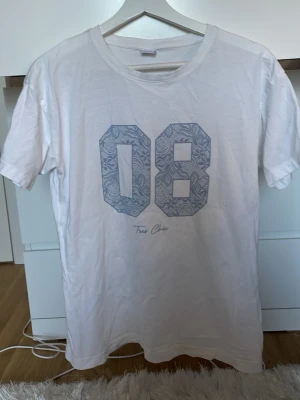 Vit t-shirt med blått tryck 08 - Säljer en vit t-shirt med stort blått tryck av siffrorna 08 på bröstet, dekorerat med ett diskret blommönster. Under siffrorna står det 'Too Chic' i skrivstil. T-shirten har rund hals och korta ärmar, perfekt för en avslappnad stil.