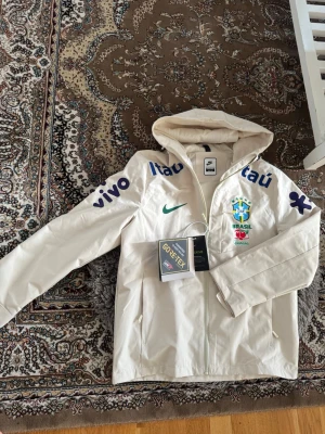 Brasil vindjacka - Snygg vit vindjacka från Nike med Brasiliens landslagslogga och sponsorer tryckta på bröstet och ärmarna. Jackan har huva, hel dragkedja och är tillverkad i ett lätt syntetmaterial. Perfekt för dig som gillar fotboll och vill ha en sportig stil. Den är storlek M men den passar XS😁🤙🏼