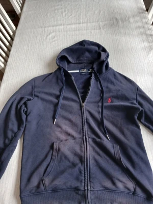 Mörkblå hoodie med dragkedja - Mörkblå hoodie från Polo Ralph Lauren med dragkedja framtill, huva med dragsko och kängurufickor. Klassisk röd logga broderad på bröstet. Perfekt för en avslappnad och stilren look. Mjuk bomullsblandning för extra komfort.