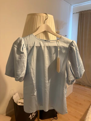 Ljusblå puffärmblus från VILA - Supersöt ljusblå blus från VILA med puffiga korta ärmar och rund halsringning. Tillverkad i ett glansigt, mjukt material som ger en elegant känsla. Perfekt att styla med jeans eller kjol för en trendig look.