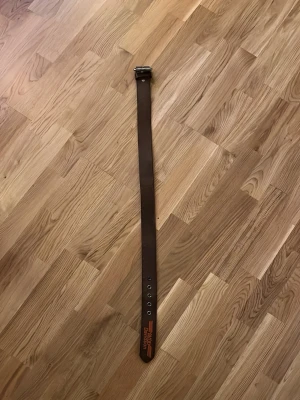 Brunt Harley Davidson skärp 94cm - Snyggt brunt skärp i skinn med klassiskt metallspänne. Skärpet har orange Harley-Davidson-logga tryckt vid änden och flera hål för justering. Perfekt accessoar för att lyfta din outfit.
