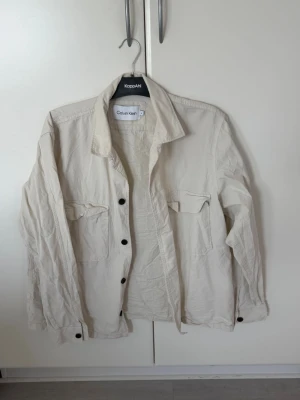Beige overshirt från Calvin Klein - Stilren beige overshirt från Calvin Klein med två bröstfickor och svarta knappar framtill. Overshirten har klassisk krage, långa ärmar med knapp vid ärmslut och en diskret logga på ärmen. Tillverkad i ett lätt bomullsmaterial, perfekt för lager på lager.