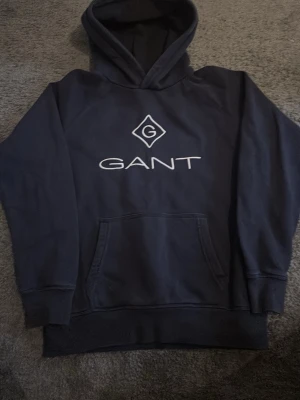 Mörkblå hoodie från GANT - Snygg mörkblå hoodie från GANT med stor logga och emblem på bröstet. Klassisk känguruficka framtill och huva med dragsko. Perfekt för en avslappnad och trendig look. Mjuk bomullsblandning och ribbade muddar vid ärmar och nederkant.