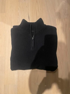 Svart stickad tröja med halvzip - Snygg svart stickad tröja med halvzip och hög krage. Tröjan har ribbstickad struktur och långa ärmar, perfekt för lager på lager. Passar till både jeans och chinos för en clean look.