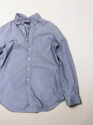 Blå skjorta från Ralph Lauren - Klassisk blå skjorta från Ralph Lauren med lång ärm och knappar framtill. Bröstficka med rosa broderad logga. Skjortan har en avslappnad passform och button-down krage. Perfekt för en stilren look.