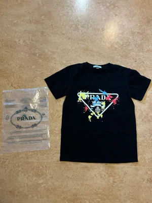 Svart t-shirt från Prada med tryck - Svart t-shirt från Prada med färgglatt tryck framtill i blått, gult och rött. Klassisk passform med rund hals och korta ärmar. Tillverkad i mjuk bomull som känns skön mot huden. Perfekt för dig som vill ha en statement-tee med exklusiv känsla. Storlek medium passar någon med small