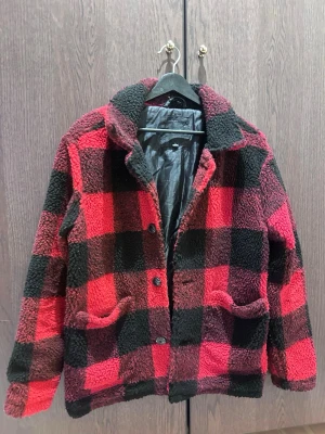 Röd och svart rutig teddyjacka - Säljer en fluffig teddyjacka i röd och svart rutigt mönster. Jackan har krage, knäppning framtill med knappar och två stora fickor. Materialet är mjukt och värmande, perfekt för kyliga dagar. Fodrad insida för extra komfort.