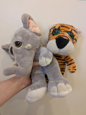 Gosedjur elefant och tiger - Två mjuka gosedjur, en grå elefant med stora öron, betar och stora bruna ögon samt en orange tiger med svarta ränder och gröna ögon. Båda är tillverkade i plysch och har gulliga detaljer som passar perfekt som kramdjur eller dekoration.