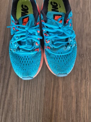Blå Nike sneakers med orange sula - Snygga turkose/ blå sneakers från Nike med ventilerande meshmaterial, svarta detaljer och orange/ turkose sula. Skorna har turkosa skosnören och orange logga på plösen. Perfekta för dig som vill ha en sportig och färgstark look.