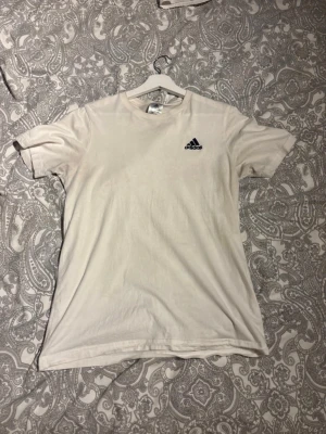 Vit t-shirt från Adidas - Klassisk vit t-shirt från Adidas med liten svart logga på bröstet. T-shirten har rund hals och korta ärmar, tillverkad i mjuk bomull för en skön känsla. Enkel och stilren design som passar till det mesta.