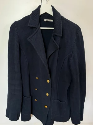 Mörkblå stickad skepparkavaj - Snygg mörkblå stickad skepparkavaj från Gina Tricot med dubbelknäppning i guld, två framfickor och klassisk krage. Perfekt för dig som vill ha en stilren och trendig look med marininspirerade detaljer.