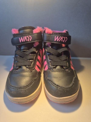 Svarta sneakers med rosa detaljer - Säljer ett par svarta sneakers med rosa fluffigt foder och rosa detaljer på sidorna. Skorna har kardborreband med texten 'WARR' och svarta skosnören. Perfekta för dig som vill sticka ut med färg och stil.