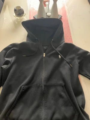 Svart hoodie från Polo Ralph Lauren - Svart hoodie med dragkedja från Polo Ralph Lauren. Klassisk design med huva och dragsnören, samt fickor framtill. Liten vit logga broderad på bröstet. Tillverkad i mjukt bomullsmaterial som är skönt att bära. Priset kan diskuteras. Knappast använd.