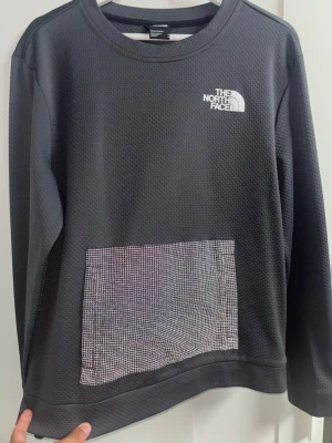 Grå The North Face sweatshirt  - Mörkgrå sweatshirt från The North Face med vit logga på bröstet och ryggen. Tröjan har en unik ficka framtill med rutigt mönster i vitt och svart reflex. Tillverkad i ett strukturerat, tekniskt material och har långa ärmar samt rund hals.