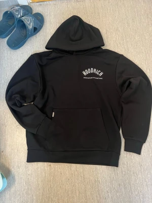 Svart hoodie från Hoodrich - Svart hoodie från Hoodrich med vit text 'HOODRICH' och 'FROM NOTHING TO SOMETHING' på bröstet och ryggen. Klassisk känguruficka framtill och huva. Perfekt för en chill och streetwear-inspirerad look.