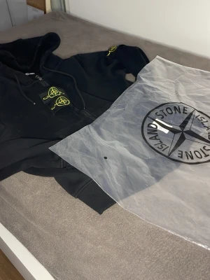 Svart hoodie från Stone Island - Säljer en svart hoodie från Stone Island med dragkedja framtill och den klassiska loggan i gult på ärmen. Hoodien har huva med dragsko och ribbade muddar. Perfekt för dig som gillar streetwear och vill ha något stilrent men ändå med edge.