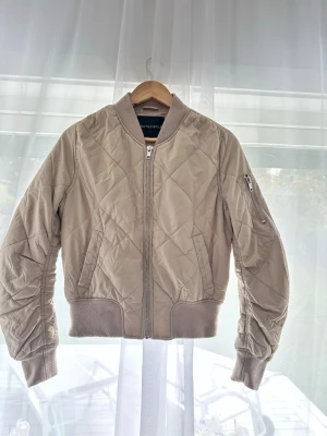 Beige quiltad bomberjacka - Snygg beige bomberjacka med quiltat mönster och ribbade muddar i ärmslut och midja. Jackan har dragkedja framtill och en ficka med dragkedja på ena ärmen. Perfekt för dig som vill ha en stilren och trendig look.