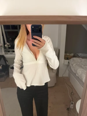 Vit v-ringad topp med lång ärm - Trendig vit topp med djup v-ringning och långa ärmar. Toppen har en lätt utsvängd passform och är tillverkad i ett ribbat material som ger en snygg struktur. Perfekt att styla med jeans eller kjol för en cool look.