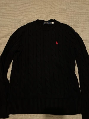 Svart kabelstickad tröja Polo Ralph Lauren - Kabelstickad svart tröja från Polo Ralph Lauren med klassisk röd logga broderad på bröstet. Tröjan har rund halsringning, långa ärmar och ribbade muddar. Perfekt för dig som gillar stilrena och tidlösa plagg.