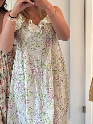 Blommig sommarklänning med volang - Söt långklänning med blommigt mönster i pastellfärger som rosa, gul och grön på vit botten. Klänningen har volangdetaljer vid axlarna och är ärmlös, perfekt för varma dagar. Tillverkad i ett lätt och luftigt material, idealisk för sommarens alla tillfällen. Den är i storlek XS men passar för S