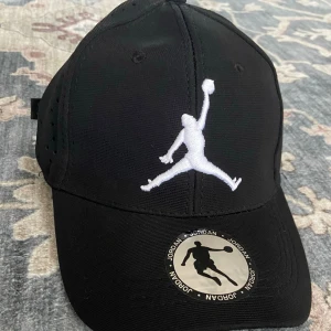 Svart keps med vit Jumpman-loga - Snygg svart keps från Jordan med den klassiska vita Jumpman-loggan broderad framtill. Kepsen har böjd skärm, ventilationshål på sidorna och justerbar rem bak för perfekt passform. Perfekt för en sportig och streetinspirerad stil.