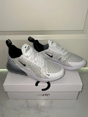 Nike Air Max 270 vita sneakers - Nike Air Max 270 sneakers i vitt med svarta detaljer och synlig luftdämpning i hälen. Ovandelen är i mesh för bra ventilation och har en modern, sportig look. Swoosh-logga på sidan och dragflik bak. Perfekta för dig som gillar stilrena och bekväma sneakers.