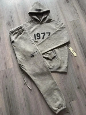 1977 essentials tracksuit  - Mysigt grått loungewear-set med hoodie och matchande byxor. Hoodien har trycket '1977' framtill och byxorna har samma tryck på benet. Setet har ribbade muddar och dragsko i midjan. Perfekt för chill dagar hemma.