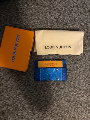 Blå Louis Vuitton korthållare - Säljer en blå korthållare från Louis Vuitton med sportigt basketplan-motiv och små spelare. Tillverkad i slittåligt läder med flera fack för kort. Kommer med original ask och dustbag. Perfekt accessoar för dig som gillar exklusiv och unik design.