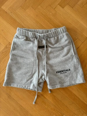 Grå shorts Essentials Fear of God - Snygga grå shorts från Essentials Fear of God med svart logga på vänster ben. Shortsen har elastisk midja med snörning och sidofickor. Tillverkade i mjuk bomull, perfekta för en avslappnad stil.