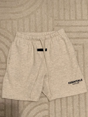 Grå Essentials Fear of God shorts - Snygga ljusgrå shorts från Essentials Fear of God med svart logga på benet. De har elastisk midja med snörning och sidofickor. Perfekta för chill dagar eller streetwear-stil.