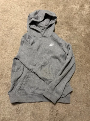 Grå hoodie från Nike med logga - Snygg grå hoodie från Nike med klassisk vit logga på bröstet. Hoodien har känguruficka framtill och mjukt, bekvämt material. Perfekt för att ha i skolan eller att träna i. Använd ca 1-2 gånger. Den är 156-170 men passar ungefär som en Xs. Skicka meddelande vid funderingar😊🙌
