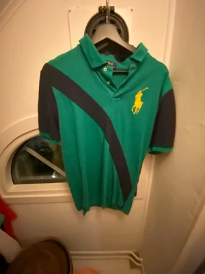 Grön piké från Polo Ralph Lauren XL - Snygg grön pikétröja från Polo Ralph Lauren i storlek XL. Tröjan har korta ärmar, mörkblå diagonal detalj och stor gul logga på bröstet. Klassisk krage med knappar och sportig look. Perfekt för dig som gillar stilrena märkeskläder.
