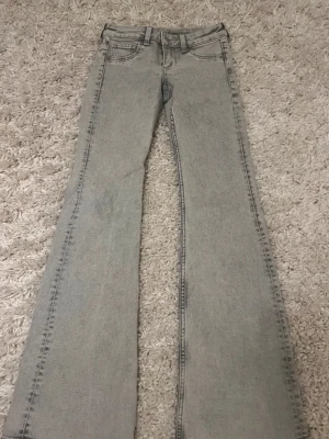 Grå bootcut jeans med låg midja - Säljer ett par ljusgrå bootcut jeans med låg midja och klassiska fem fickor. Jeansen har utsvängda ben och en snygg vintagekänsla. Perfekta till sneakers eller boots för en trendig look.