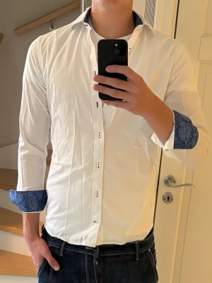 John Henric Skjorta   - Ursnygg skjorta från John Henric med oerhört snygga och stilrena detaljer! En väldigt exklusiv skjorta som är i otroligt bra kvalitet och har en mycket behaglig passform. Skjortans vita färg gifter sig perfekt med de blåa detaljerna!