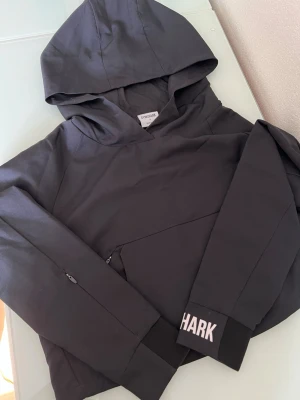 Svart hoodie från Gymshark - OANVÄND. Snygg svart hoodie från Gymshark med huva och dragkedjeficka på sidan. Ärmslutet har en cool detalj med Gymshark-loggan i vitt. Perfekt för gymmet eller chill. Materialet känns mjukt och stretchigt för maximal komfort.