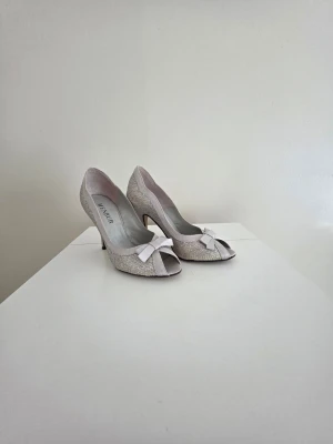 Glittriga peep toe pumps med rosett - Snygga pumps i glittrigt silvermaterial med öppen tå och söt rosett framtill. Skorna har smal klack och elegant form som ger en lyxig känsla. Perfekta för dig som vill sticka ut med lite extra bling.