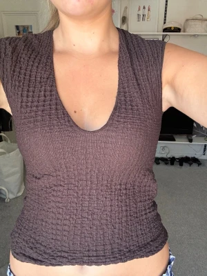 Brun ribbad ärmlös topp - Säljer en brun ärmlös topp med ribbad struktur och djup v-ringning. Toppen har en tight passform och är perfekt för dig som gillar en enkel men ändå trendig stil. Materialet känns mjukt och stretchigt, vilket gör den bekväm att bära hela dagen.