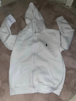 Vit hoodie med dragkedja och huva - Säljer en vit hoodie med dragkedja och huva. Hoodien har två fickor framtill och en liten broderad logga på bröstet. Perfekt för en avslappnad och stilren look. Materialet känns mjukt och bekvämt.