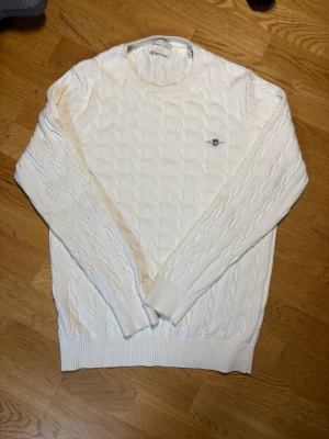Vit kabelstickad gant tröja - Kabelstickad vit tröja med rund halsringning och ribbade muddar vid ärmslut och nederkant. Gant logga på bröstet. Klassisk och stilren design som passar till mycket. Materialet är  mjukt och bekvämt. Vid frågor är det bara att skicka meddelande.