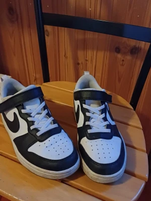 Svarta och vita sneakers med kardborre - Snygga sneakers i svart och vitt med klassisk design och kardborreband. Skorna har vit bas med svarta detaljer och en tydlig logga på sidan. Ovandelen är i syntetmaterial och de har vita skosnören samt perforerad tå för extra ventilation.
