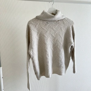 Beige stickad polotröja - Säljer en mysig vit stickad polotröja. Den har ett fint flätmönster och en hög polokrage som håller dig varm under kalla dagar. Tröjan är långärmad och perfekt för höst och vinter. Observera att det finns ett hål på framsidan som behöver lagas. Perfekt för dig som gillar att pyssla eller vill ha en tröja att mysa i hemma.