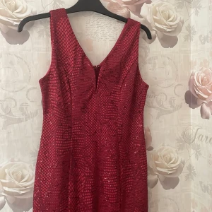 Röd glittrig långklänning - Säljer en superfin röd långklänning med glittrigt mönster. Klänningen har en v-ringad hals och är ärmlös, perfekt för fest eller bal. Materialet känns som polyester och den har en tight passning som framhäver figuren. Klänningen är i nyskick och redo för att användas på nästa stora event! Använd en gång. Köparen står för frakten!